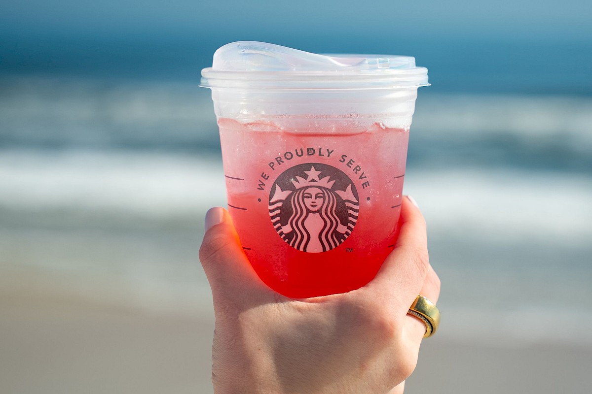 A hand holds a Starbucks cup filled with a pink drink in front of a beach setting.