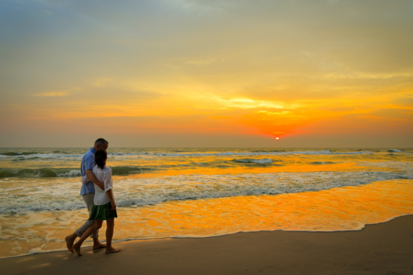 A couple is walking along a beach during a beautiful sunset, with waves gently crashing on the shore.