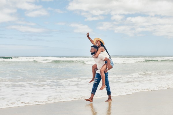 A couple enjoys a playful moment on the beach, with one giving the other a piggyback ride under a partly cloudy sky.