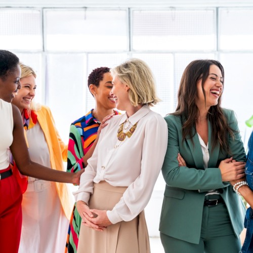Six women are happily interacting and sharing a laugh in a bright, modern indoor space.