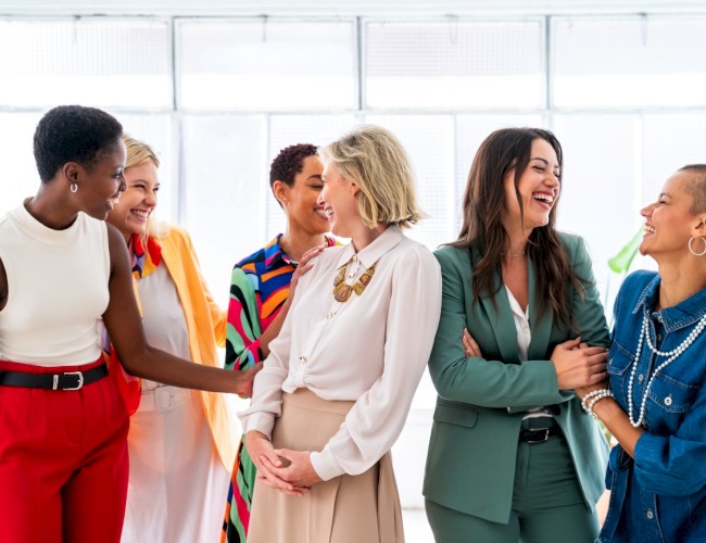 Six women are happily interacting and sharing a laugh in a bright, modern indoor space.