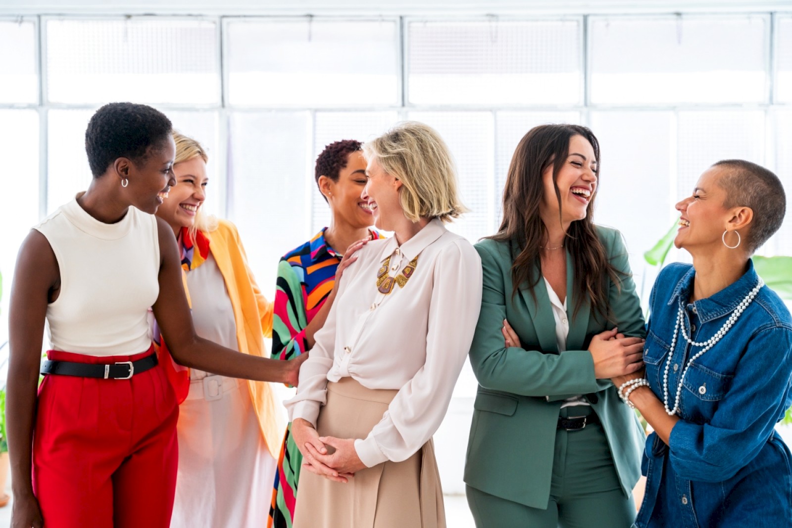 Six diverse women are happily chatting and laughing together in a bright, modern office.