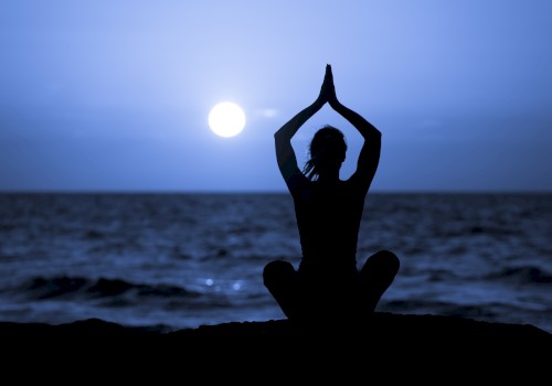 A person sits in a meditative pose by the sea at twilight, with hands raised, silhouetted against a setting or rising sun over the water.