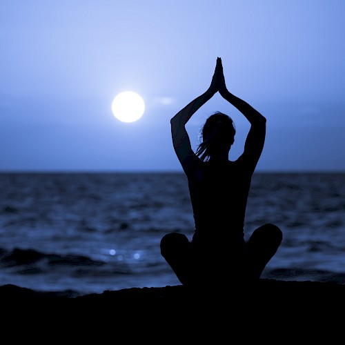 A person sits in a meditative pose by the sea at twilight, with hands raised, silhouetted against a setting or rising sun over the water.