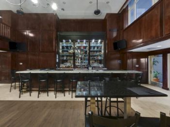 This image displays an upscale bar area with high wooden panels, a stocked bar shelf, a long counter with high chairs, and modern lighting.