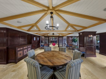 The image shows a dining area with a round table and six striped chairs, wooden beams on the ceiling, and large windows providing natural light.