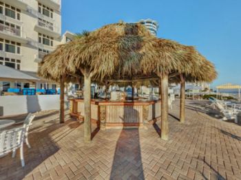 The image shows an outdoor tiki bar with a thatched roof, surrounded by table seating. It's located near a multi-story building and a coastal area.