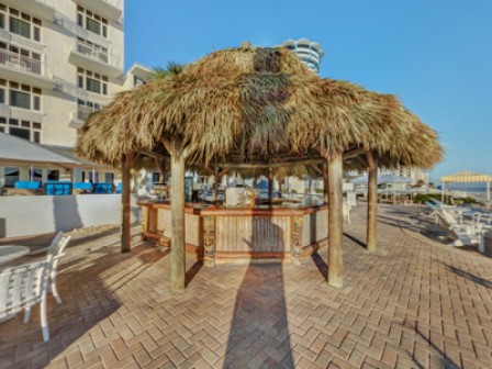 The image shows an outdoor tiki bar with a thatched roof, surrounded by table seating. It's located near a multi-story building and a coastal area.