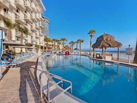 The image shows a hotel pool area with lounge chairs, palm trees, a tiki hut, and an ocean view in the background, on a sunny day.