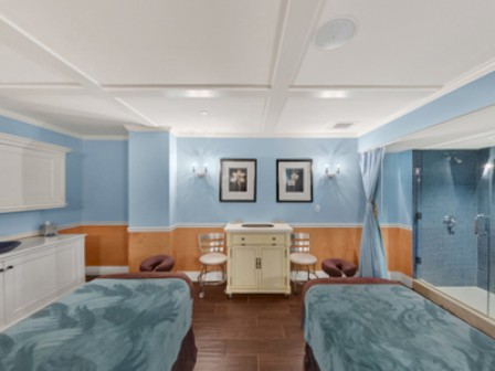 A serene room with two massage tables, framed art, white cabinetry, blue walls, and a glass-enclosed shower area, suitable for a spa or wellness center.