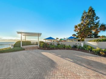 The image shows a paved walkway near the ocean, with a pergola, landscaped greenery, and blue umbrellas near a seating area, under a clear blue sky.