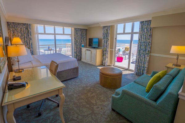 A bright hotel room with a view of the beach, featuring a bed, TV, desk, sofa, and a balcony overlooking the ocean.