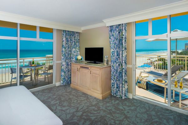 A hotel room with large windows, ocean view, TV, bed, floral curtains, and a balcony with outdoor seating and umbrella overlooking the ocean.