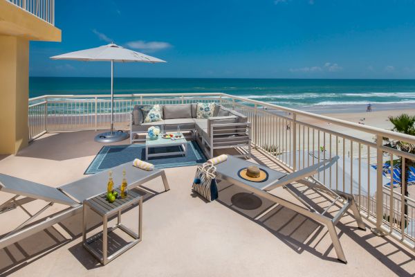 A balcony with lounge chairs, a table, drinks, a bag, and a hat overlooks the beach and ocean, featuring an umbrella and seating area.