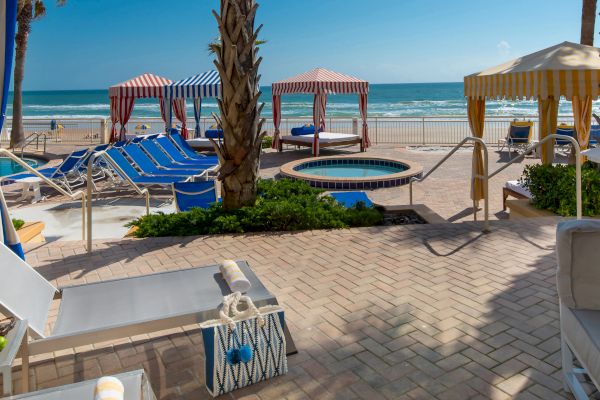 A beachside area with cabanas, lounge chairs, a small pool, palm trees, and a view of the ocean under a clear blue sky.