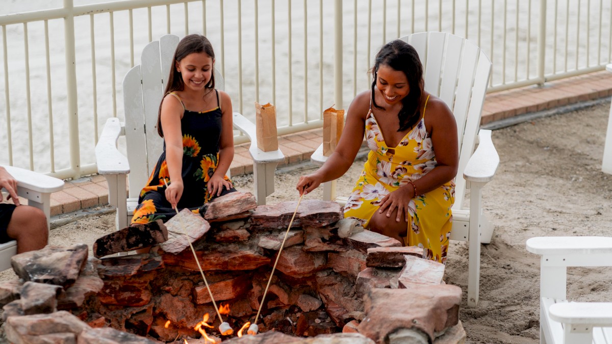 Two people are by a fire pit on a sandy area, sitting in chairs, and roasting marshmallows with sticks.