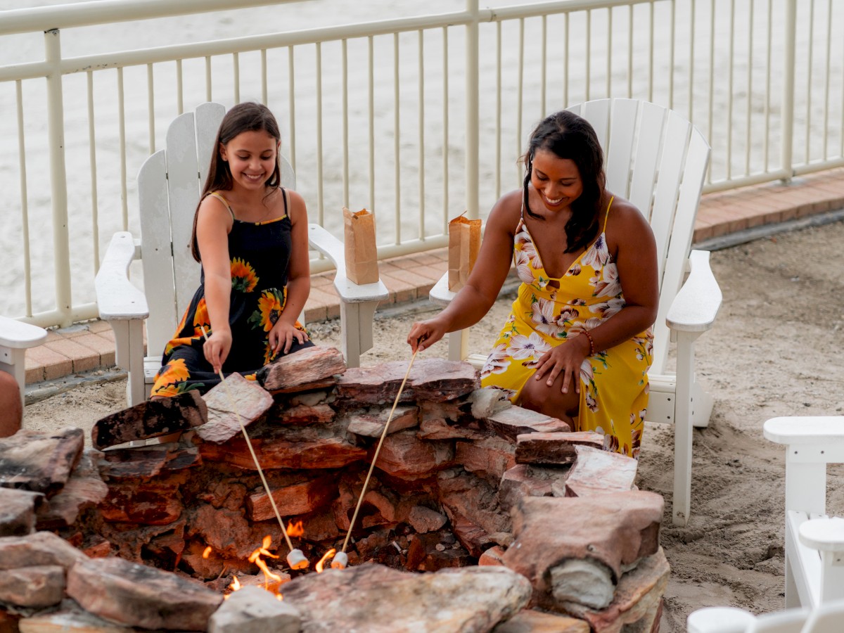 Two people are by a fire pit on a sandy area, sitting in chairs, and roasting marshmallows with sticks.