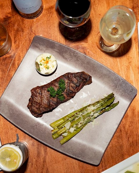 A meal with steak, asparagus, and sauces, alongside wine, lemon water, and a dessert shot on a wooden table.