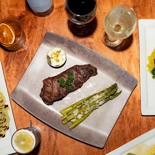 A meal with steak, asparagus, and sauces, alongside wine, lemon water, and a dessert shot on a wooden table.