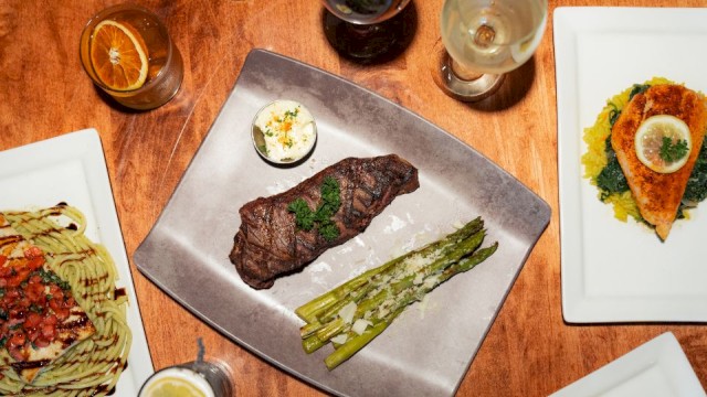 A meal with steak, asparagus, and sauces, alongside wine, lemon water, and a dessert shot on a wooden table.
