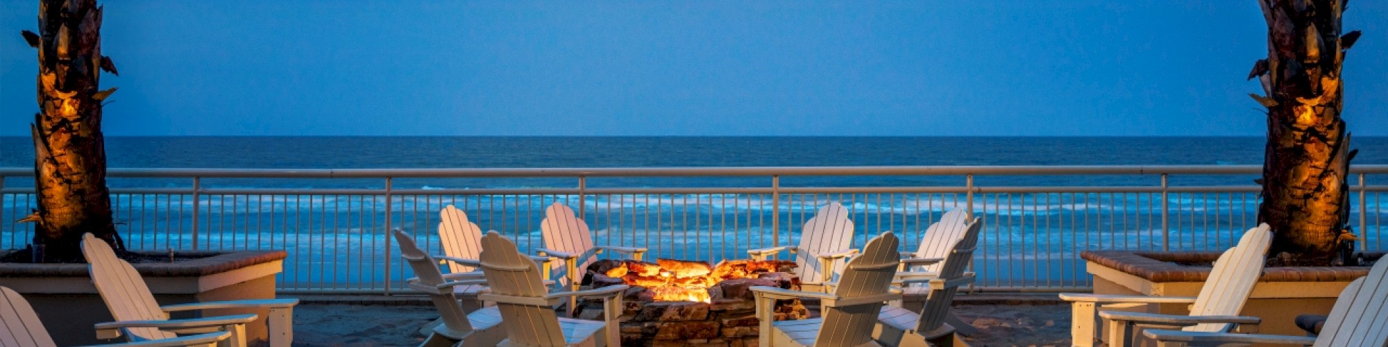 A seaside view with Adirondack chairs arranged around a lit fire pit, framed by palm trees, under a blue evening sky.