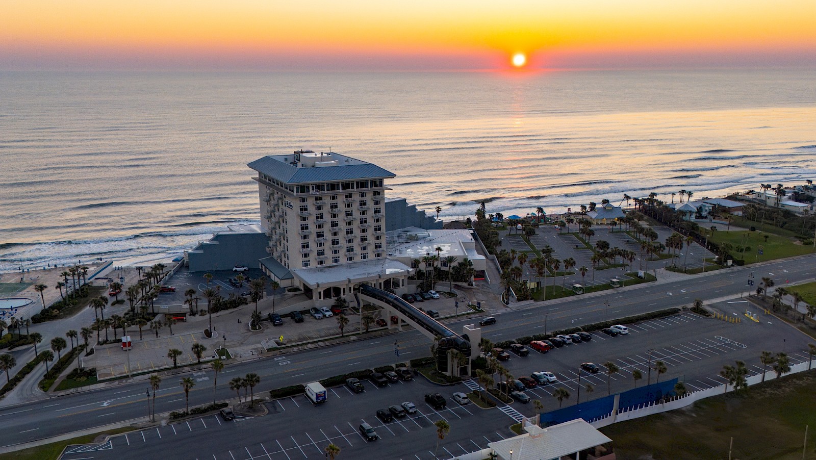 Aerial view of a beachfront hotel complex at sunset, with a tall central building, palm trees, a highway, and a parking lot by the ocean.