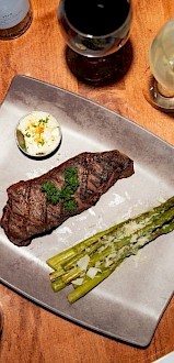 A meal with steak, asparagus, and side dishes, accompanied by wine, on a wooden table.