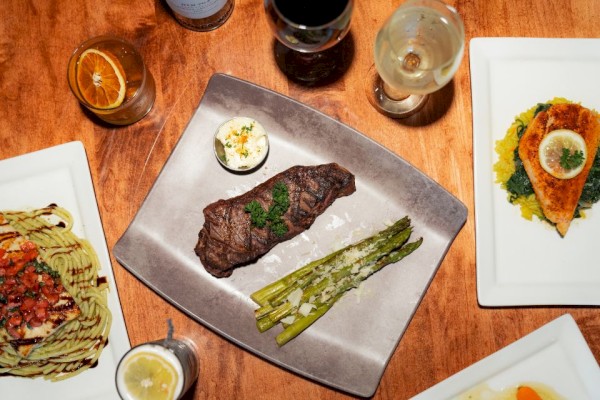 A steak with grilled asparagus, two glasses of wine, a cocktail, and several plates of pasta are on a wooden table.