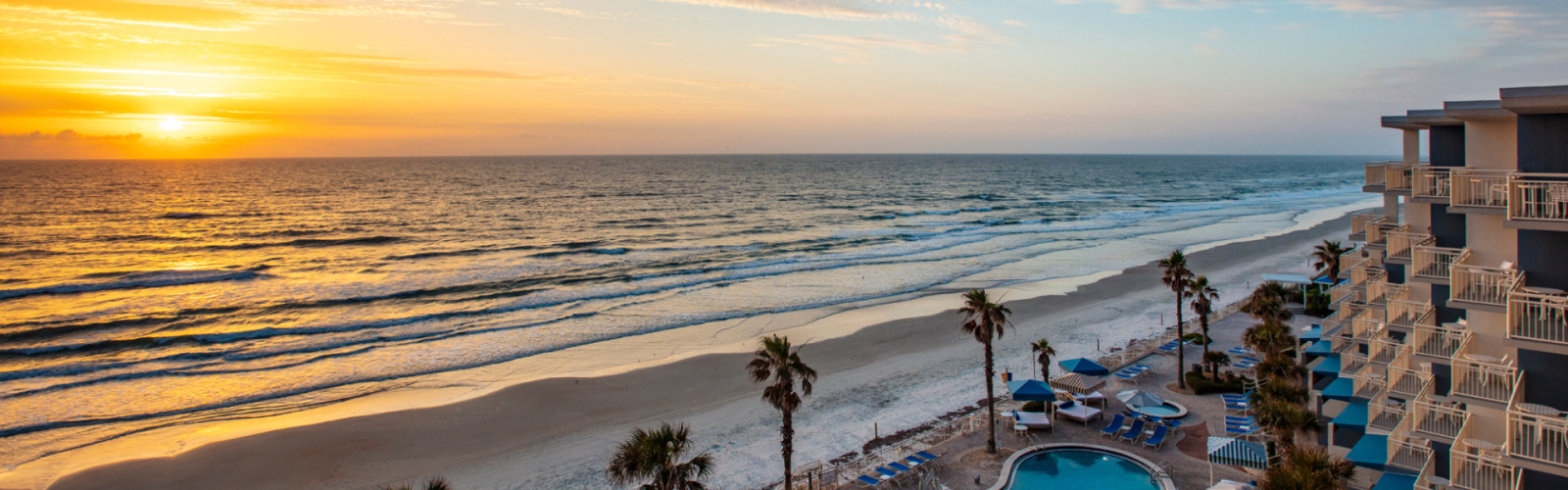 An ocean sunrise view with a sandy beach, palm trees, a pool, and a multi-story building with balconies.