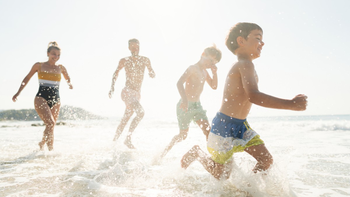 A family is running and playing in the shallow water at the beach on a sunny day, enjoying the waves and the pleasant weather, ending the sentence.