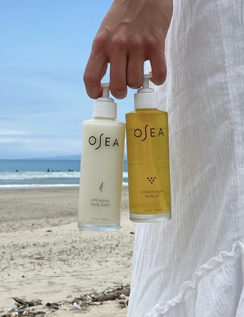 The image features a person holding two bottles of Osea skincare products, with a beach and ocean in the background.