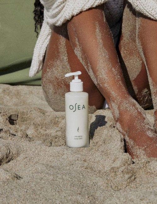 A person sitting on sand at the beach with a bottle of OSEA anti-aging body balm nearby.
