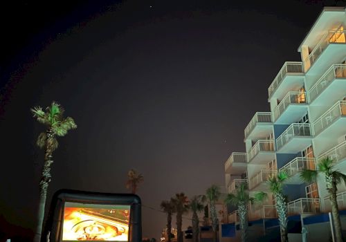 This image shows a nighttime scene with a pool, palm trees, a large screen displaying an image, and a multi-story building on the right.