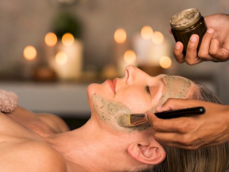 A woman receives a facial mask treatment while relaxing during a spa session, with candles in the background creating a calming atmosphere.