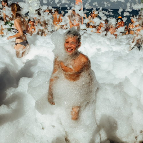 A person enjoys a foam party, surrounded by bubbles and people in swimwear, with a joyful crowd in the background under clear skies.