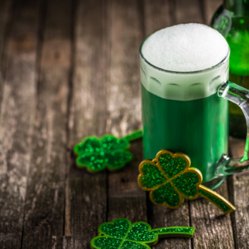 A mug of green beer with a foamy head is on a wooden surface, accompanied by glittery green shamrock decorations, suggesting a St. Patrick's Day theme.