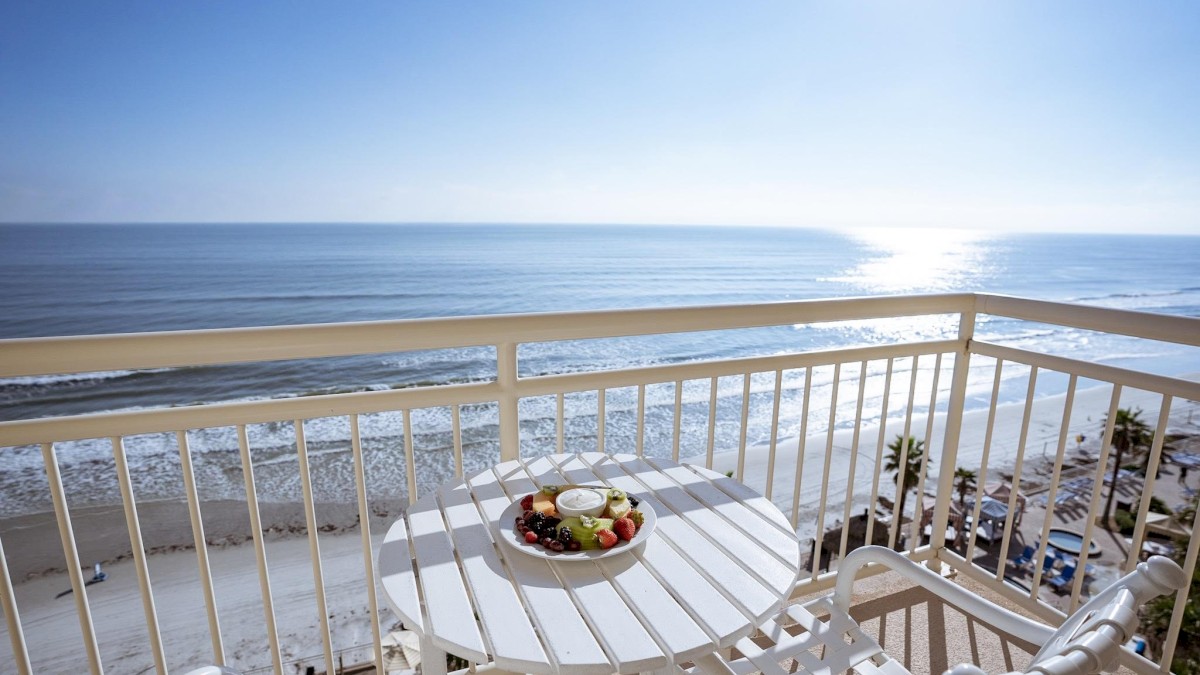 A balcony with a white table and chairs overlooking a beach and ocean, with a bowl of fruit on the table under a bright, clear sky ending the sentence.