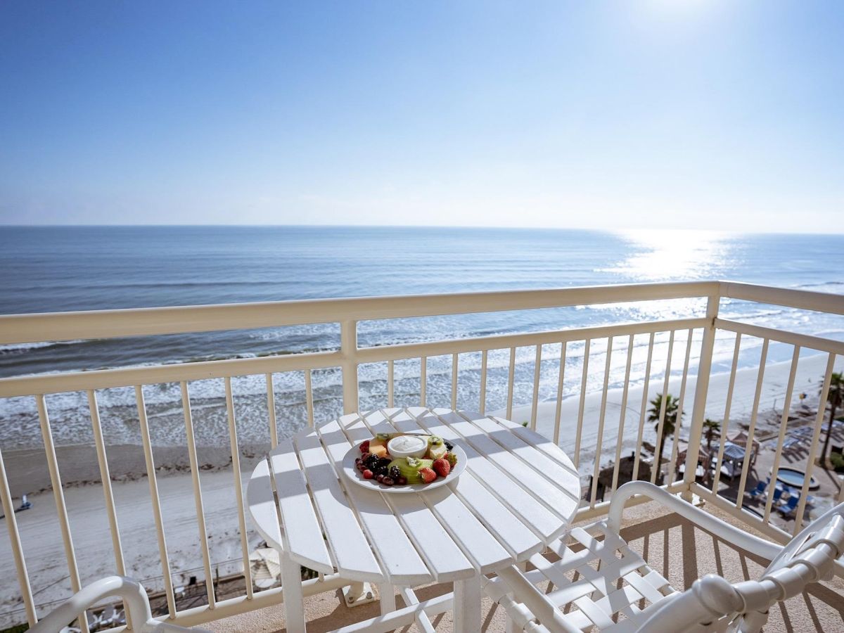 A balcony with a white table and chairs overlooking a beach and ocean, with a bowl of fruit on the table under a bright, clear sky ending the sentence.