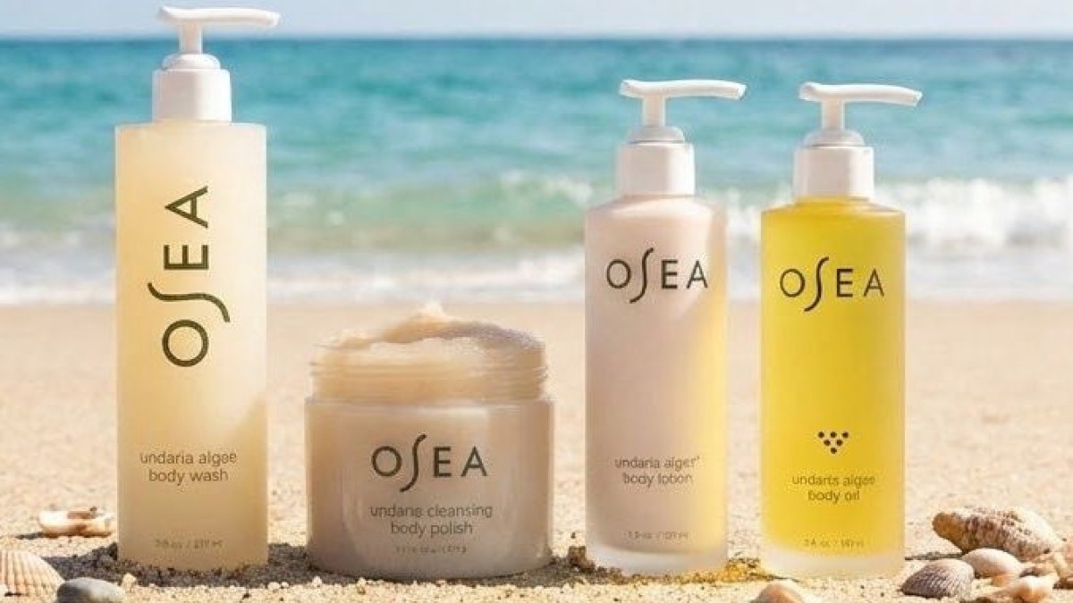 The image shows four beach-themed skincare products on sand, with the ocean and sky in the background.