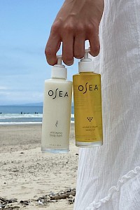 A hand holds two bottles of Osea skincare products, standing on a beach with the ocean and sky in the background.