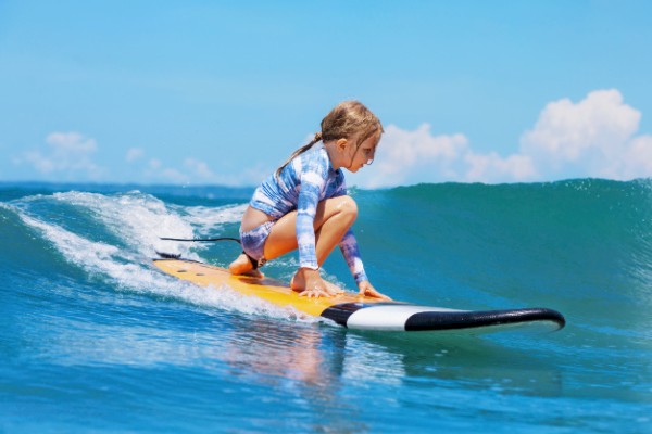 A young girl is surfing on a yellow surfboard, crouched low, wearing a long-sleeved bathing suit, and riding a small wave on a sunny day.