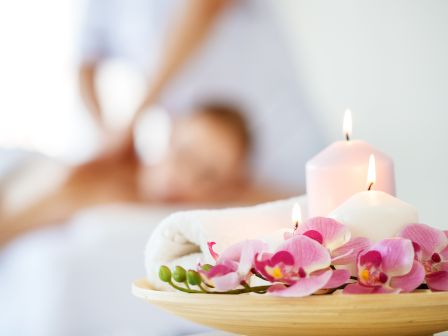 A spa scene with orchids, lit candles, and a towel in the foreground, while a person is receiving a massage in the blurred background.
