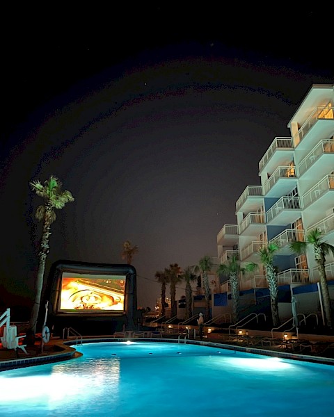 The image shows a night scene at a poolside area with palm trees, a large inflatable screen, and a multi-story hotel building.