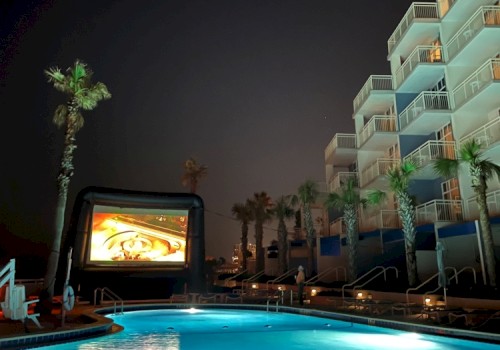 The image shows a night scene at a poolside area with palm trees, a large inflatable screen, and a multi-story hotel building.