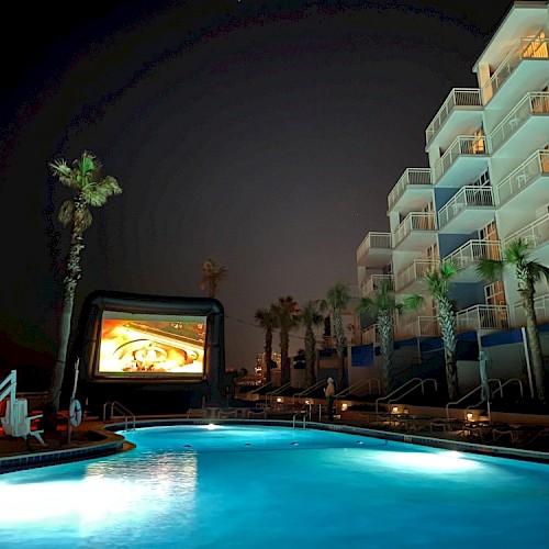 The image shows a night scene at a poolside area with palm trees, a large inflatable screen, and a multi-story hotel building.