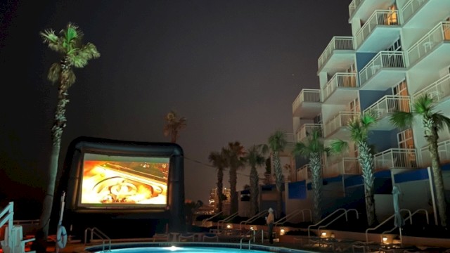 The image shows a night scene at a poolside area with palm trees, a large inflatable screen, and a multi-story hotel building.