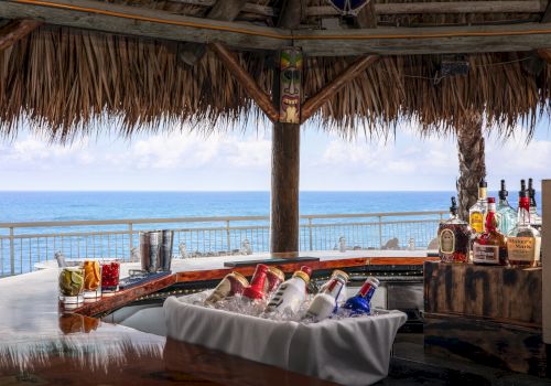 A tropical bar with various drinks, spirits, and beers on ice, overlooking a scenic ocean view under a thatched roof.