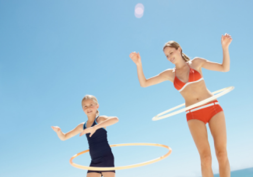 Two people are hula hooping on a sunny day, wearing swimsuits, with a clear blue sky and ocean in the background.