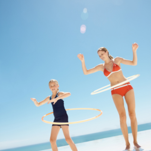 Two people are hula hooping on a sunny day, wearing swimsuits, with a clear blue sky and ocean in the background.