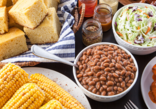 The image showcases a spread of cornbread, coleslaw, baked beans, corn on the cob, ribs, and sauces on a table, epitomizing a classic barbecue meal.